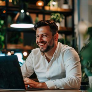 A man with dark hair and a beard, wearing a white shirt, smiles while working on a laptop at an SEO Company NYC office, warmly lit and modern, with plants and shelves in the background.