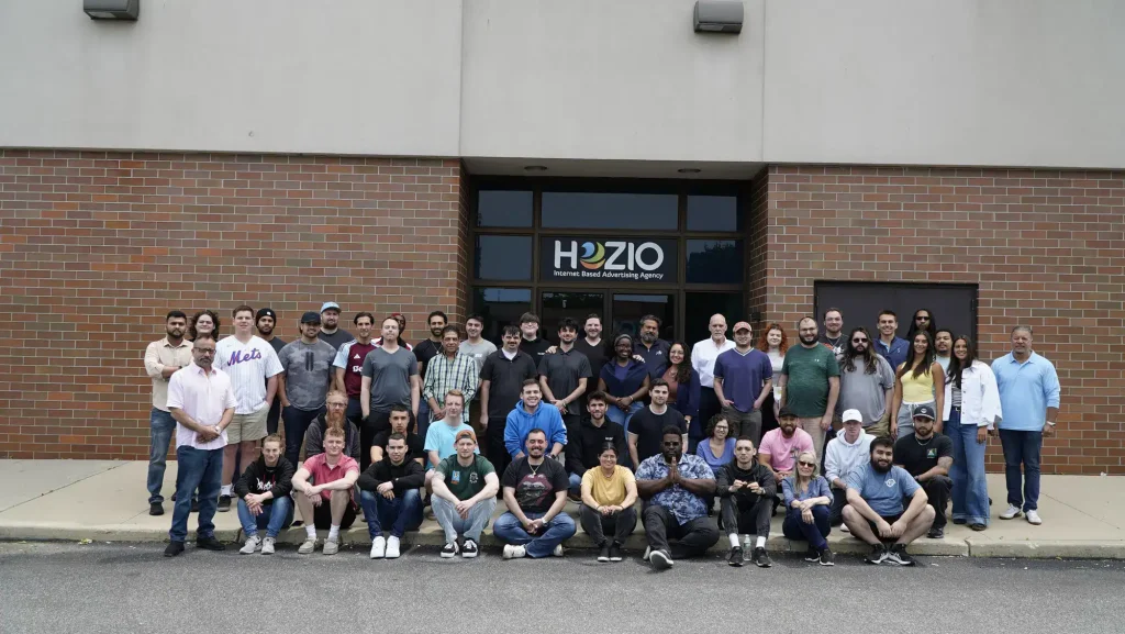 A large group of people pose for a group photo outside a brick building with a sign that reads "Hozio Internet Based Advertising Agency," a leading SEO Company NYC. Most are standing, some squatting in front, all smiling at the camera.