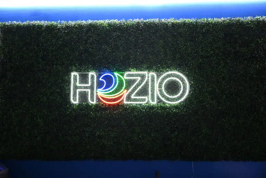 A neon sign reading "HOZIO," with colorful curved lines inside the "O," glows against a green hedge wall with blue lighting above—perfect décor for an innovative SEO Company NYC or a Search Engine Optimization Company Long Island.