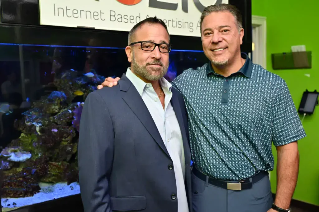 Two men standing side by side and smiling for the camera in NY; one in a suit and glasses, the other in a patterned polo. They pose before a large fish tank and a sign reading "Internet Based Advertising," representing a leading SEO Company NYC.