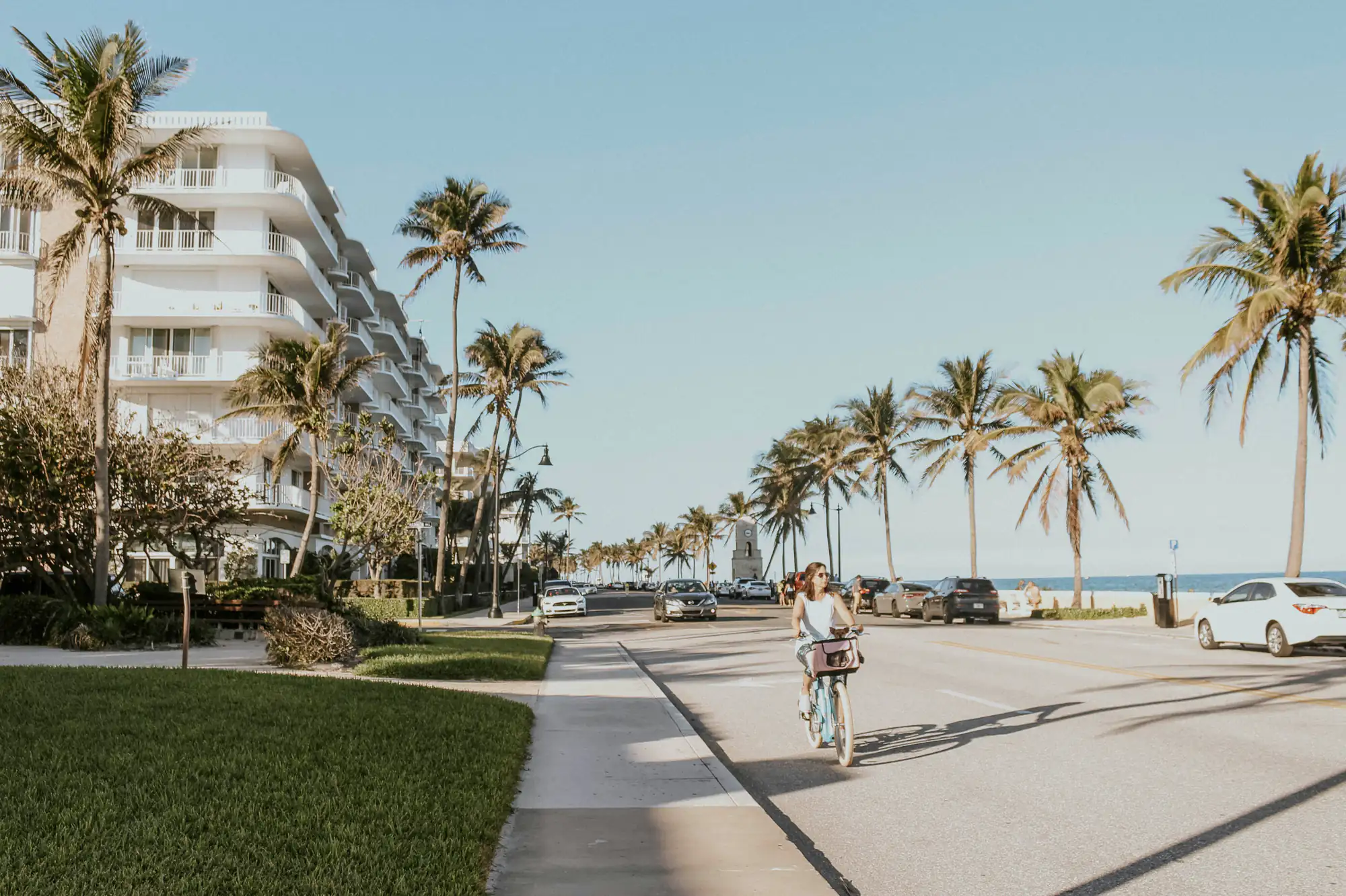 A woman rides a bicycle past palm trees and parked cars by a modern SEO Company NYC office in sunlight.