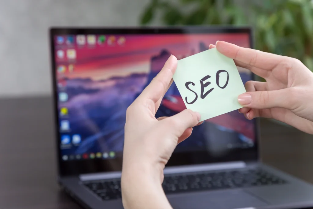 Hands display a sticky note reading “SEO” in front of a laptop, ideal for an SEO Company NYC visual.